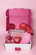 Pink gift box with various items including cosy socks, candles, eye mask, hair scrunchie, bath bomb and cosmetic bag
