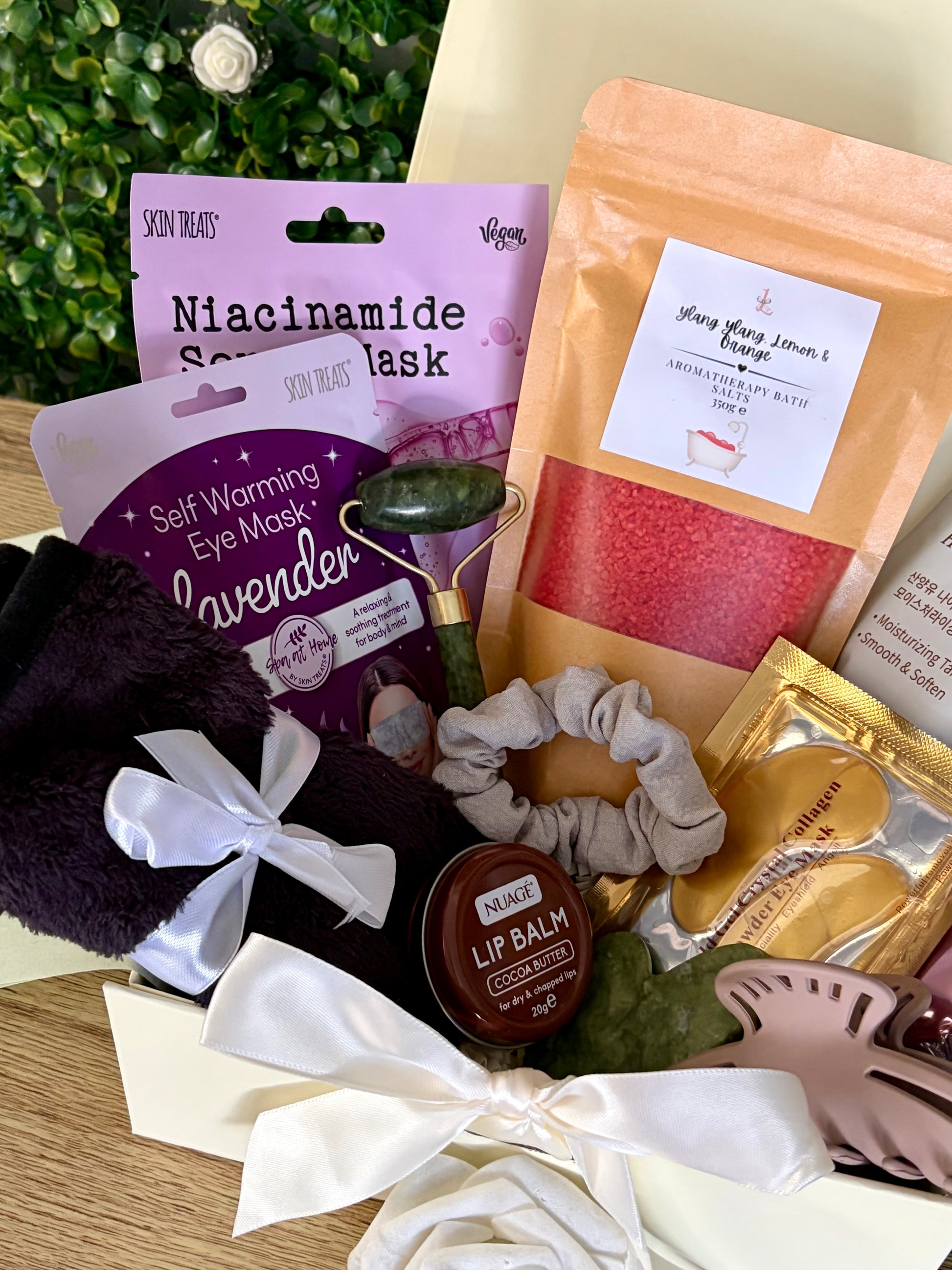 A curated self-care gift set arranged in a box, featuring a lavender Skincare Treats niacinamide mask pack, a purple lavender eye mask pouch, a peach-colored bath salt pouch labeled 'Ylang ylang, Lemon & orange', a grey scrunchie, a beige hair clip, a gold packet, a brown lip balm,black face cloth tied together with white satin ribbon.