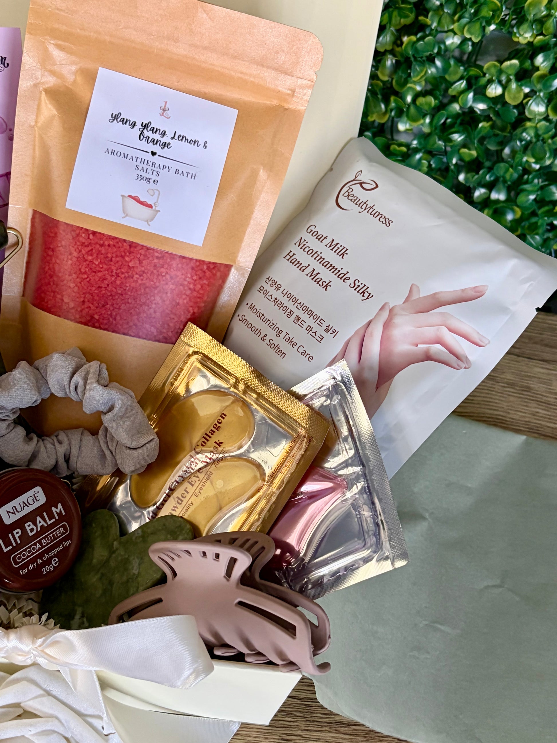 A curated gift set of self-care and beauty items arranged in a box, including a pink bath salt pouch, a white hand mask, gold and pink eye patches, a gray scrunchie, a pink hair clip, and a lip balm.
