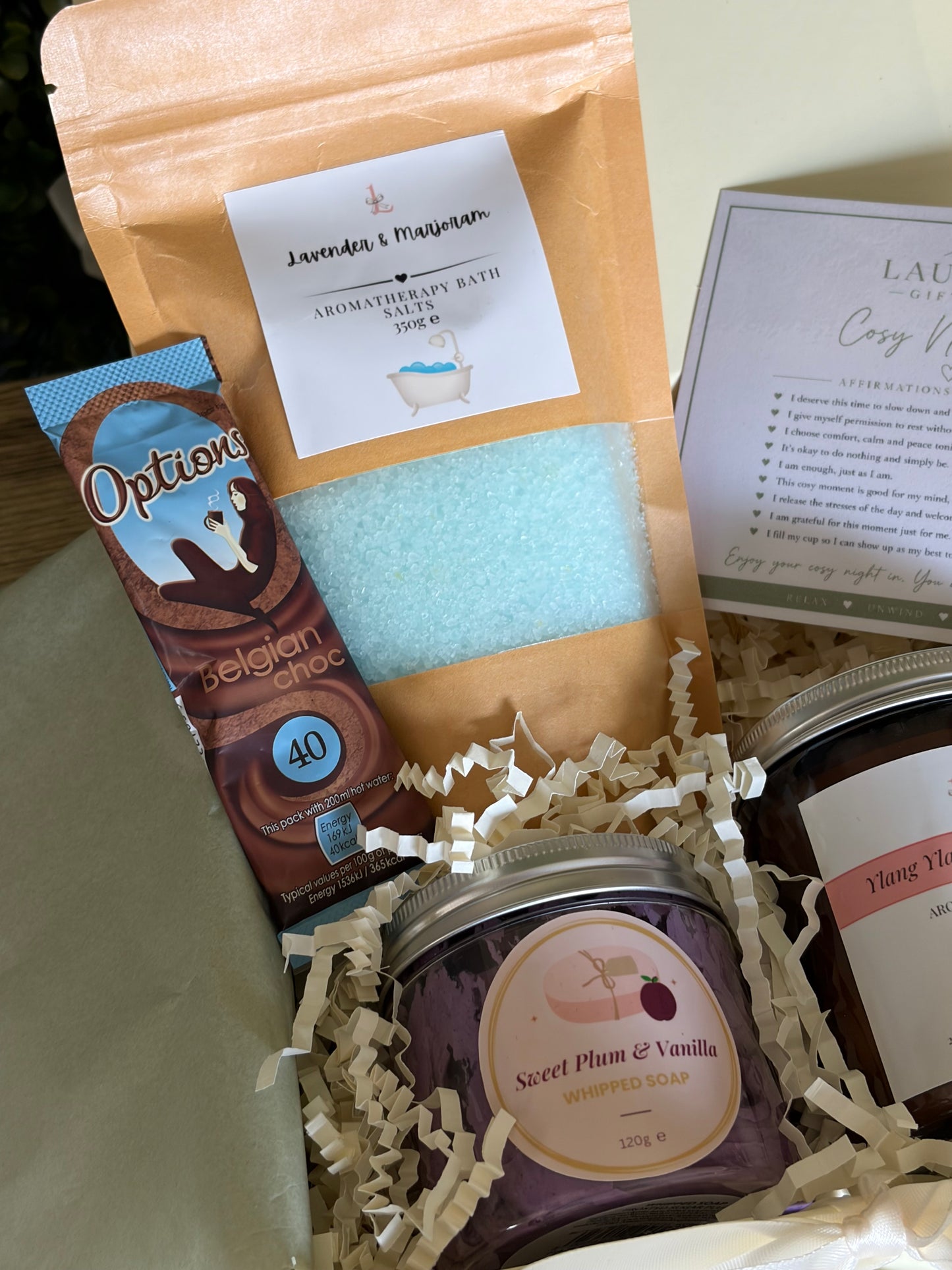 A curated gift box set containing a brown kraft pouch of lavender and marjoram bath salts,, a hot chocolate sachet, whipped soap, arranged in cream crinkle paper.
