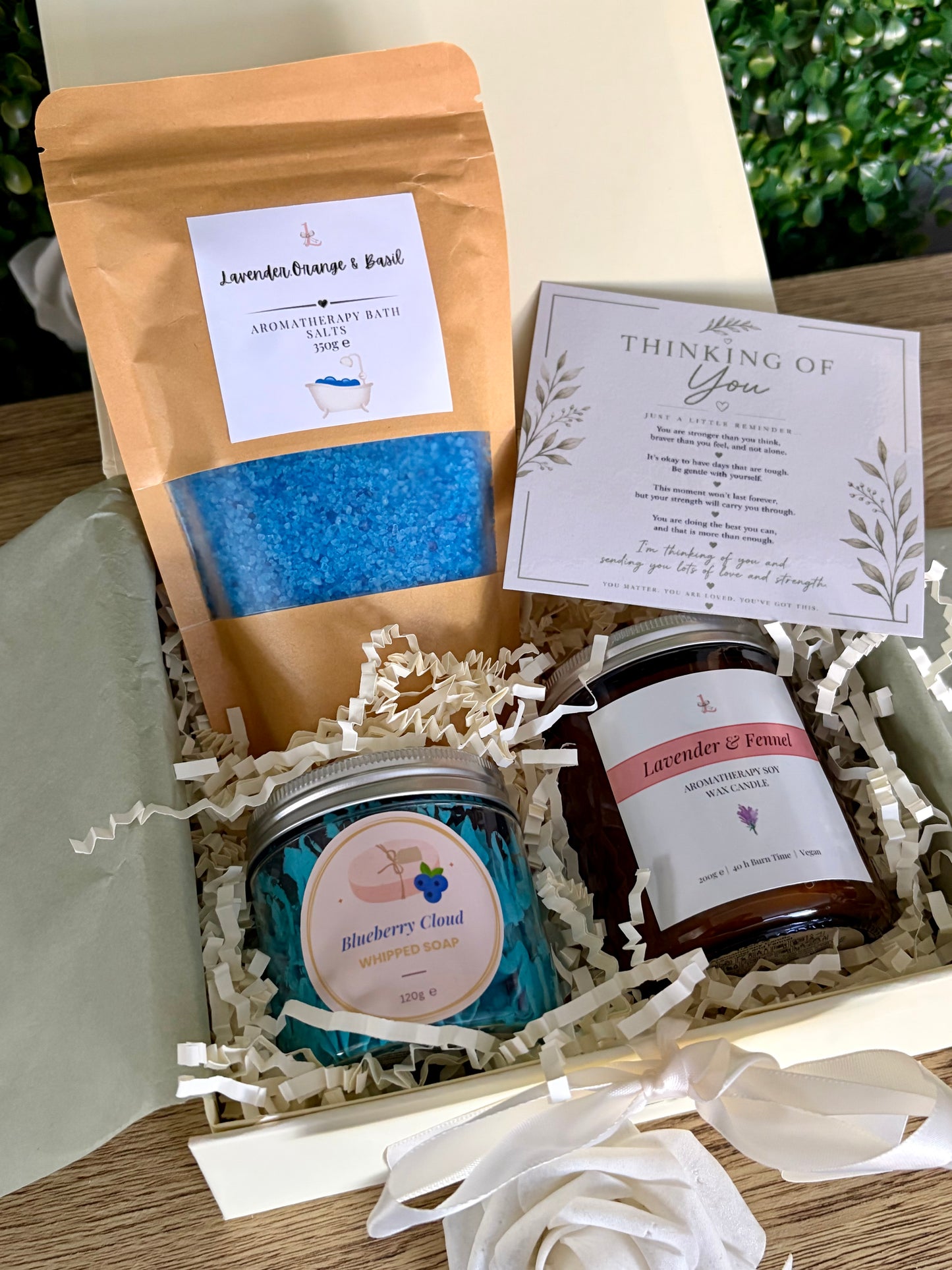 A gift box containing a lavender and orange & basil bath salts pouch, a blue 'Blueberry Cloud' whipped soap, a brown 'Lavender & Fennel' candle, and a 'Thinking of You' card, arranged in white paper shred.