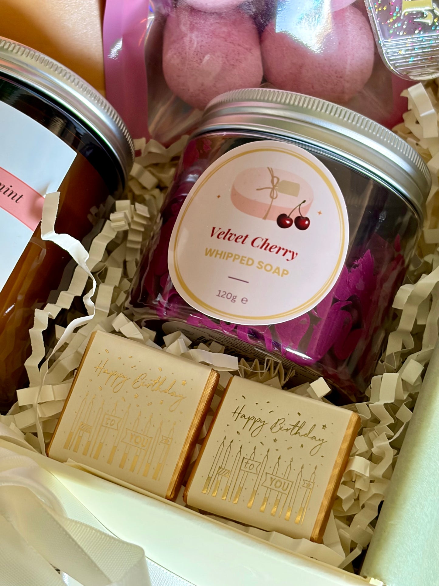 A gift set in a cream-colored box filled with shredded paper, featuring a jar of 'Velvet Cherry Whipped Soap',  , two gold-embossed 'Happy Birthday' chocolate bars, and pink bath bombs.