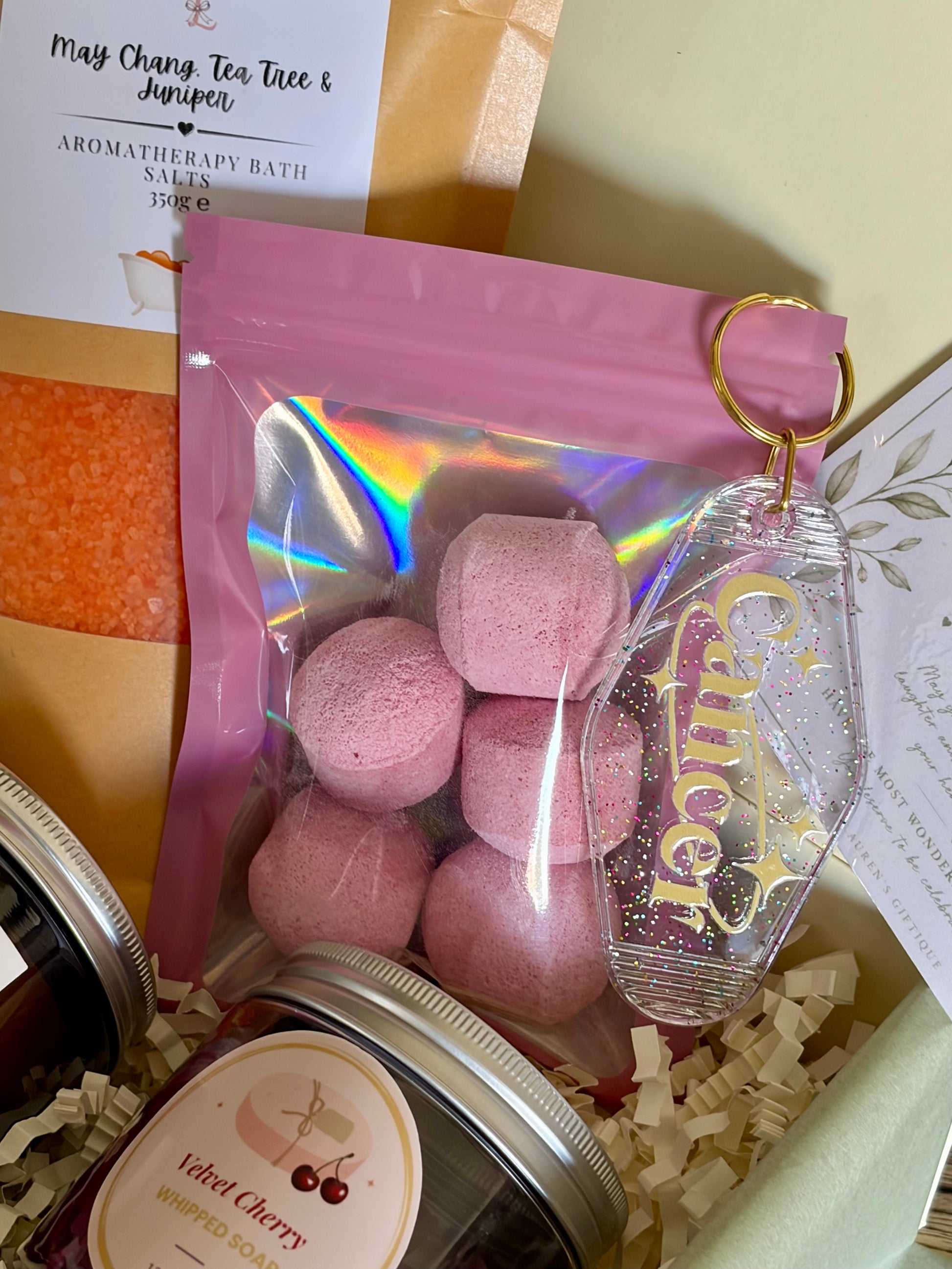 A gift box arrangement featuring a pink resealable pouch with pink bath bombs inside, a clear glittery keychain tag with gold lettering, and part of a jar labeled velvet cherry whipped soap, set in cream paper filler with care package cards.