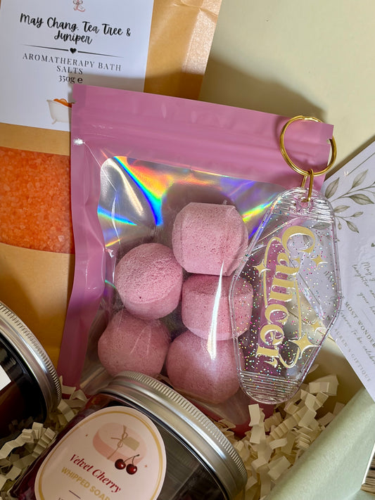 A gift box arrangement featuring a pink resealable pouch with pink bath bombs inside, a clear glittery keychain tag with gold lettering, and part of a jar labeled velvet cherry whipped soap, set in cream paper filler with care package cards.