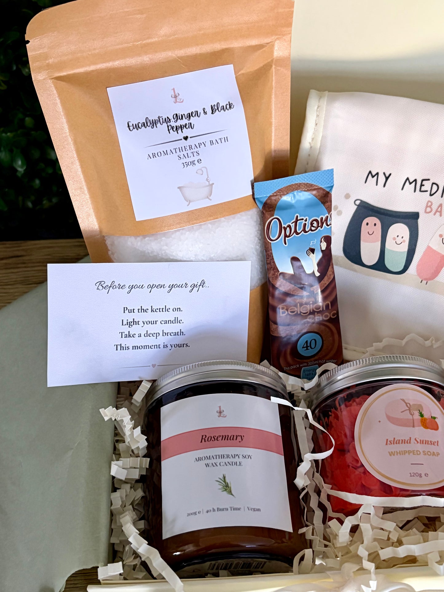 A curated spa gift set arranged in a box with shredded paper filler, including a kraft pouch labeled 'Eucalyptus, Ginger & Black Pepper' bath potion, a white card with instructions, a hot chocholate sachet, a cream pouch with cartoon characters and 'MY MEDICATION BAG', glass jar candle and whipped soap.