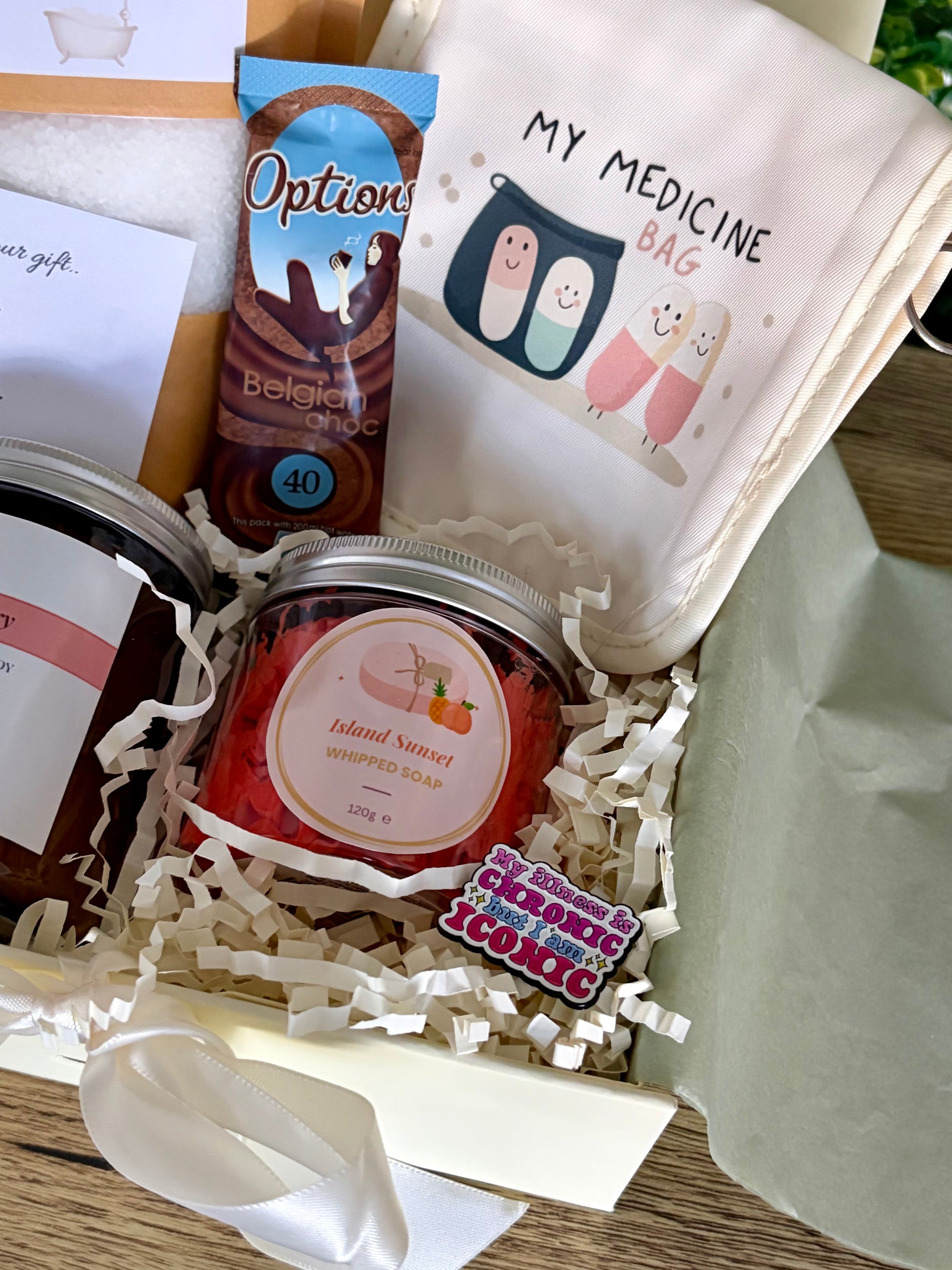 A curated gift box filled with assorted items, including a jar of Island Sunset whipped soap, a hot chocolate sachet, a cream bag printed with 'MY MEDICINE BAG' and cartoon pills, and a small badge