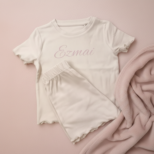 A two-piece children's pajama set in a soft light beige color, featuring a short-sleeve top with 'Ezmai' printed in pink script and matching shorts with scalloped edges,