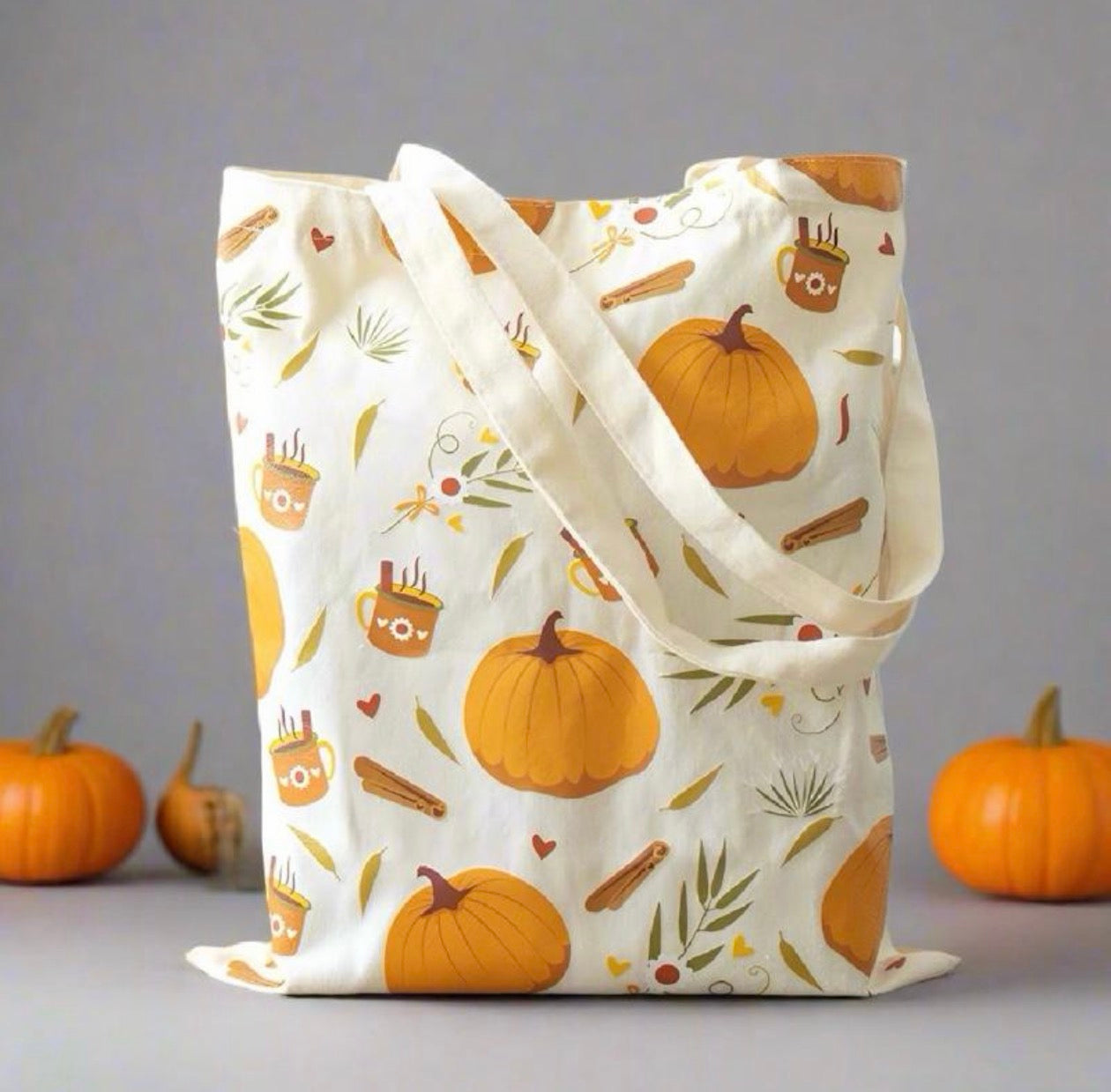 An off-white canvas tote bag with a cozy autumn pattern of orange pumpkins, cinnamon sticks, acorns, and small hearts, styled with three small pumpkins on a gray background.