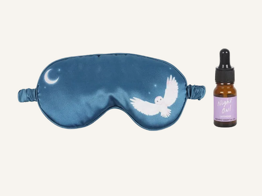 navy blue satin sleep mask with crescent moon and owl design and bottle of lavender essential oils on a ivory background.