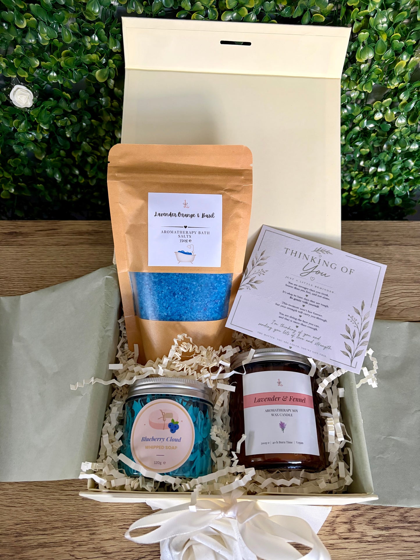 A curated gift box set containing a blue bath salt pouch, a  'Blueberry Cloud' whipped soap, a brown 'Lavender and Fennel' candle jar, a cream gift card, , all arranged in a white box with shredded paper on a wooden table with a green leafy background.
