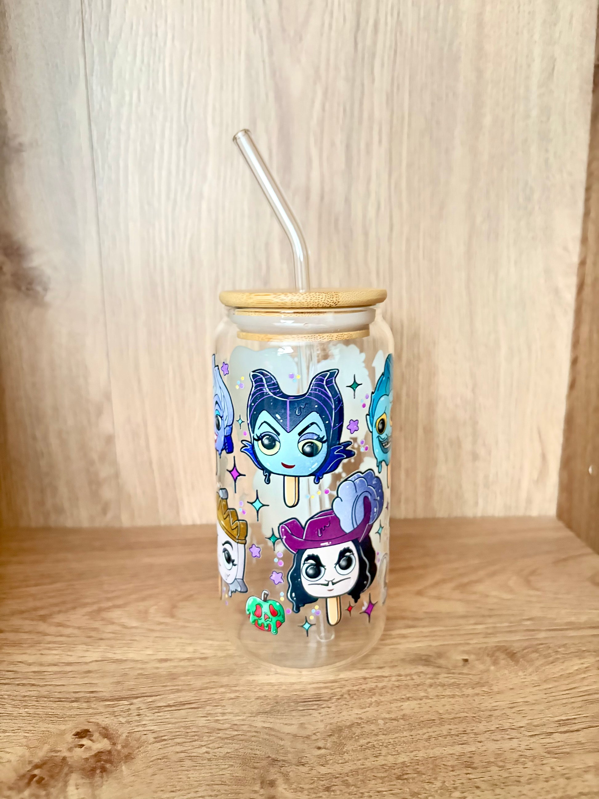 A clear glass tumbler with a bamboo lid and clear glass straw, decorated with colorful cartoon character illustrations, set on a wooden surface against a wooden background.
