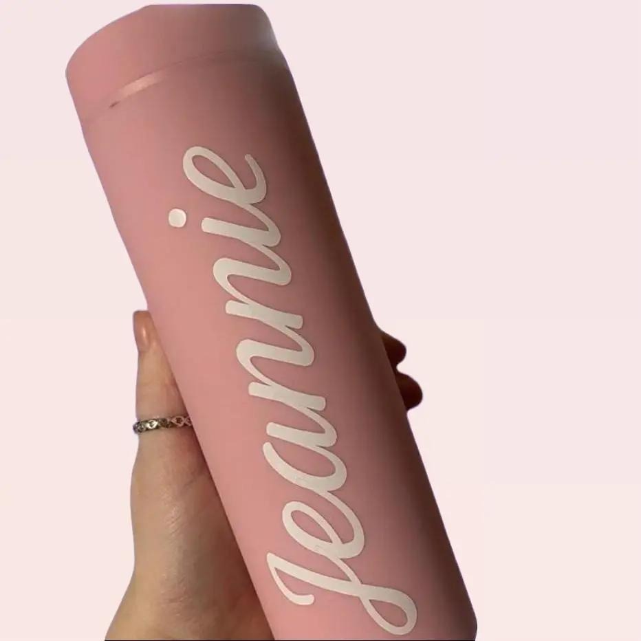 Pink tumbler container with 'jeannie' written on it, held by a hand against a light pink background.
