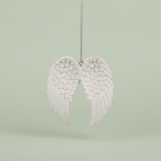 A white angel wing hanging ornament suspended by a silver string against a soft, light sage green background