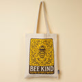 Beige tote bag with a bee and floral design, featuring the text 'BEE KIND', on a yellow background.