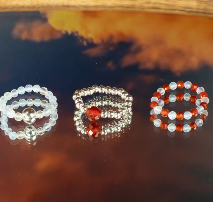Three beaded rings with red and white stones on a reflective surface