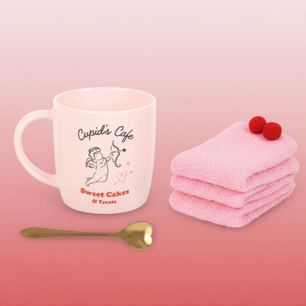 Pink mug with 'Cupid's Cafe' text, pink socks with red pom-poms, and gold heart-shaped spoon on a pink background