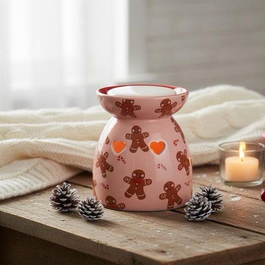 A pink ceramic oil burner candle warmer decorated with brown gingerbread men and orange heart cutouts, styled on a rustic wooden table with pinecones and a lit candle in a glass holder.