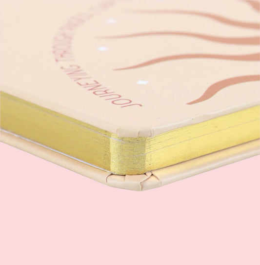 Close-up of a notebook with gold binding and 'Surrounding' branding on a pink background