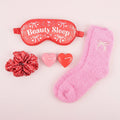 Red 'Beauty Sleep' eye mask, pink socks, Red scrunchie, and heart-shaped candles on a pink background