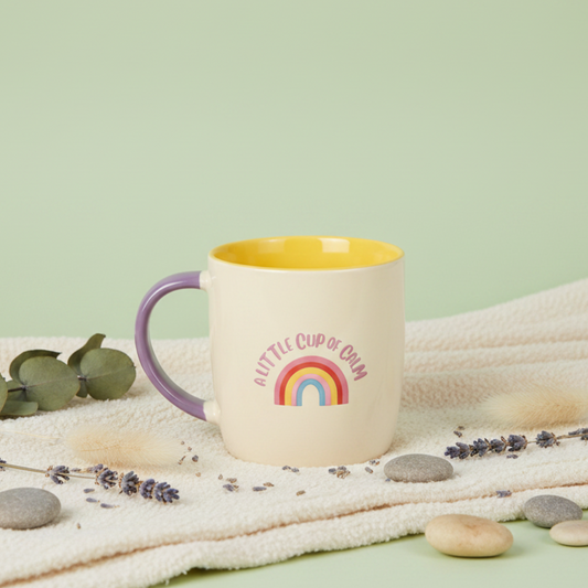 a little cup of calm mug with rainbow design on a sage green background with ivory blanket and stones