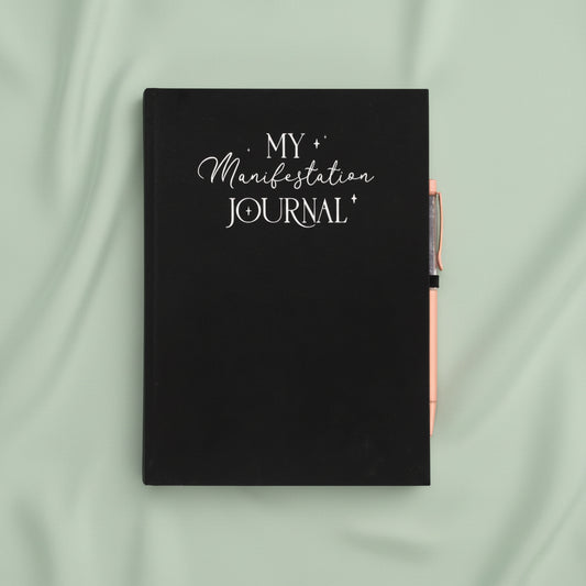 A black hardcover journal with 'MY Manifestation JOURNAL' printed in white script on the front cover, shown closed with a rose gold pen tucked into the side pocket, against a plain sage green background.