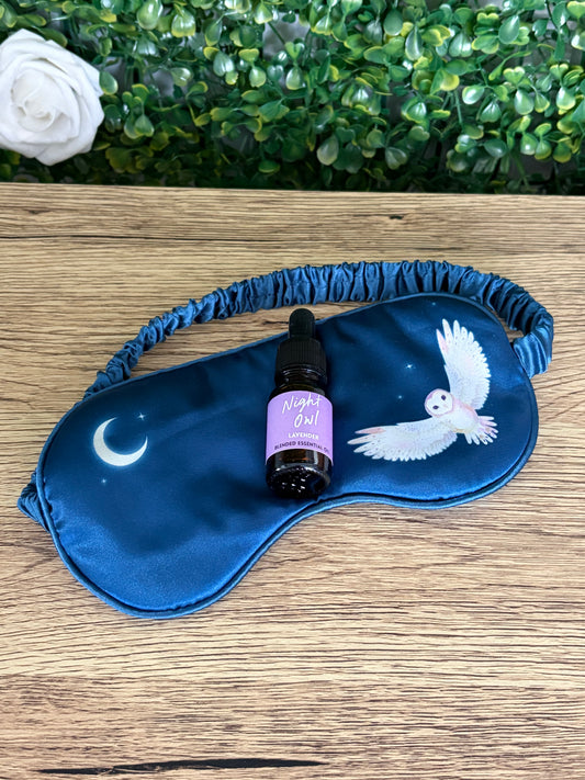 A blue satin sleep mask with a crescent moon and white owl design, displayed on a wooden surface with a small lavender dropper bottle in the center. A green leafy hedge and a white rose are visible in the background.