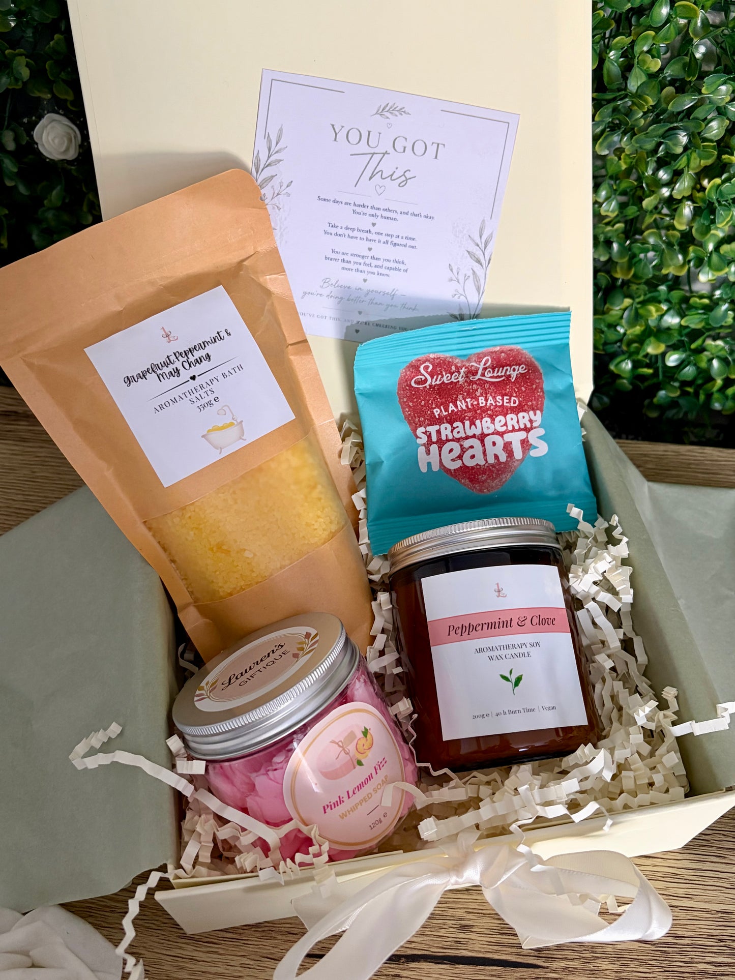 A curated gift box containing bath potion in kraft bag, sweets, whipped soap and aromatherapy candle jar on a wooden surface in ivory coloured gift box.