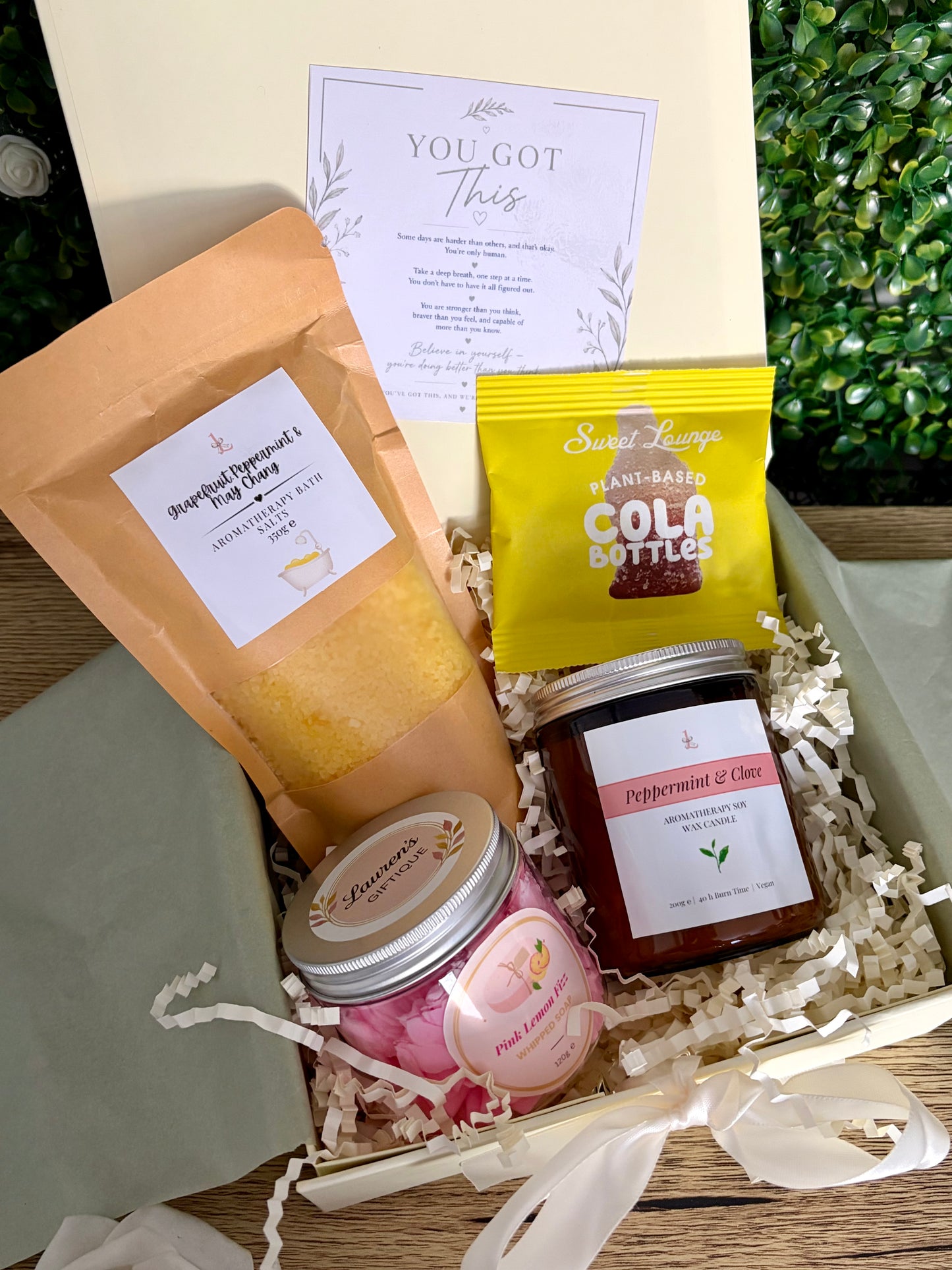 A curated gift set containing pouch of bath salts, cola bottle sweets, pink whipped soap in jar and glass candle, on a ivory filled gift box.