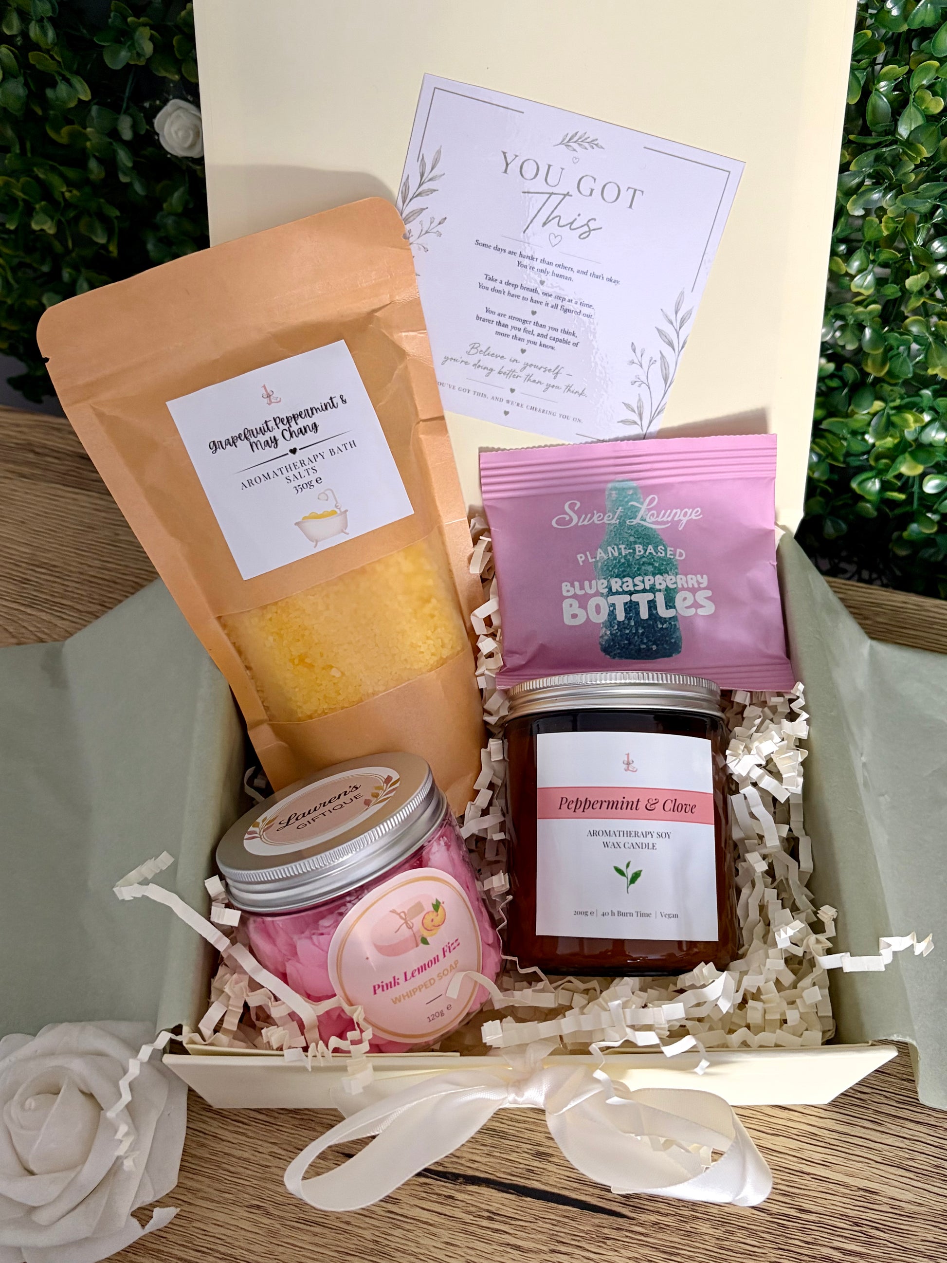 A curated gift box of bath salts, pink lemon fizz whipped soap, aromatherapy amber jar candle and packet of sweets in a vory gift box with filler and ribbon.