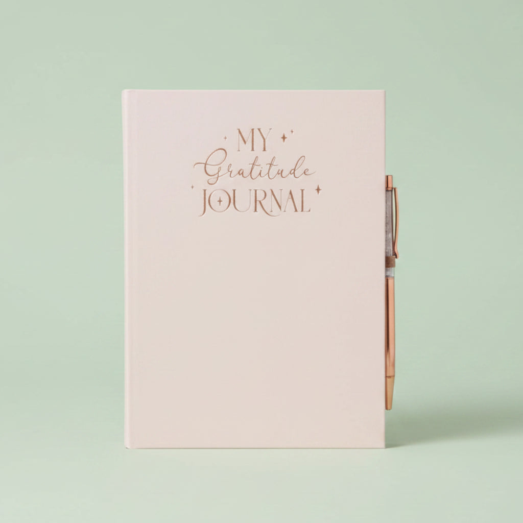 A pale blush pink hardcover journal with rose gold foil lettering that reads 'MY Gratitude JOURNAL' on the front cover, shown with a matching rose gold pen attached on the right side.