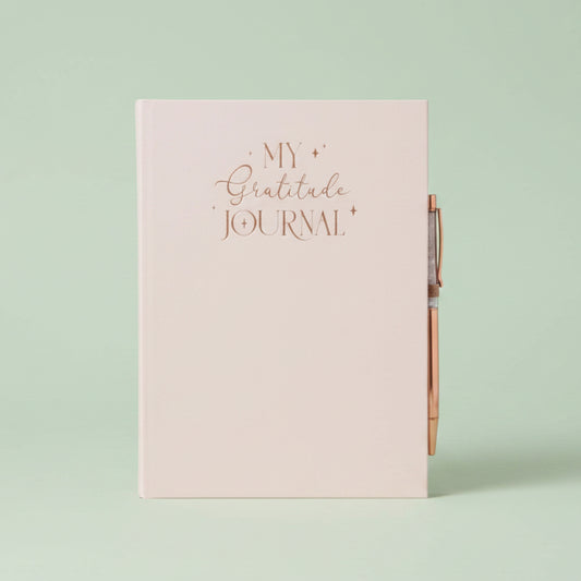 A pale blush pink hardcover journal with rose gold foil lettering that reads 'MY Gratitude JOURNAL' on the front cover, shown with a matching rose gold pen attached on the right side.