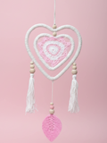 Heart-shaped crocheted dreamcatcher with tassels and beads against a pink background