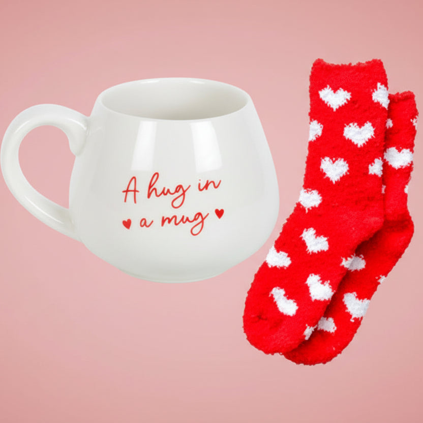 White mug with 'A hug in a mug' text and red socks with white hearts on a light background