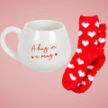White mug with 'A hug in a mug' text and red socks with white hearts on a light background