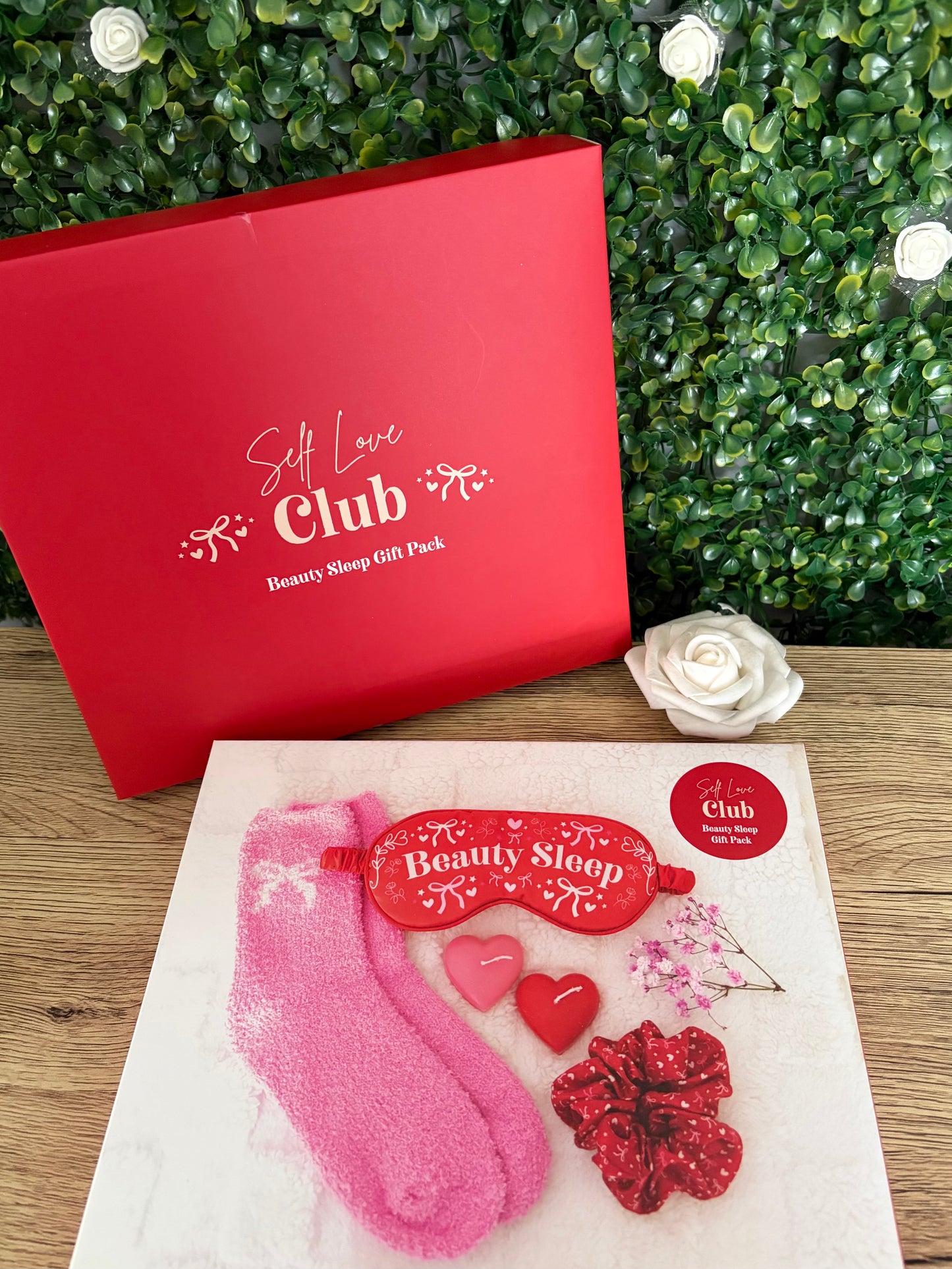 Self love club beauty sleep gift pack in a red and white box with red satin eye mask, pink fluffy socks, heart candles and hair scrunchie on a wooden surface with green leafy background