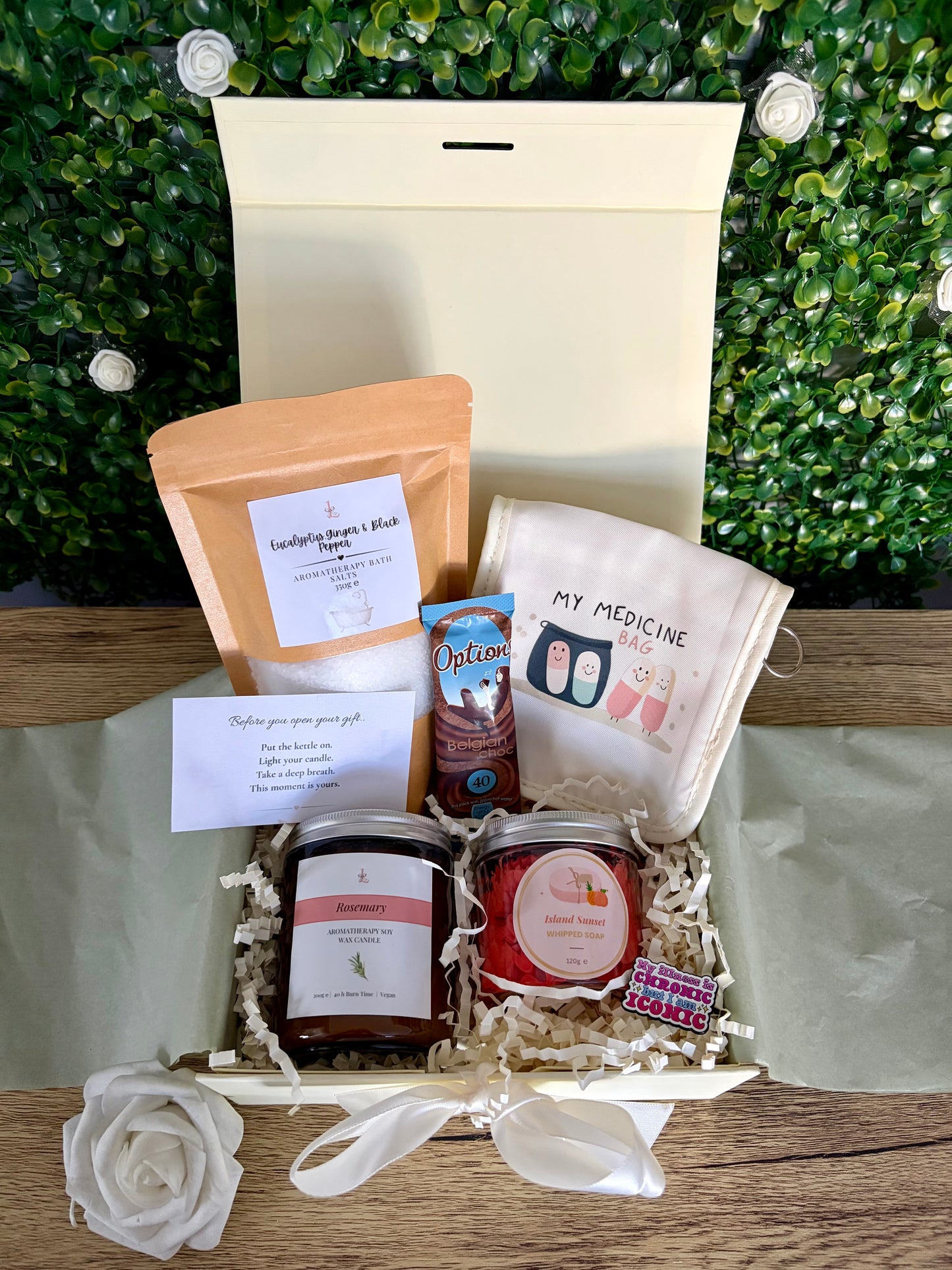 A curated gift box arrangement on a wooden surface with a green leafy backdrop. The box contains a cream medicine pouch, a kraft pouch, a  candle, a jar of whipped soap, a card, and shredded paper filler.