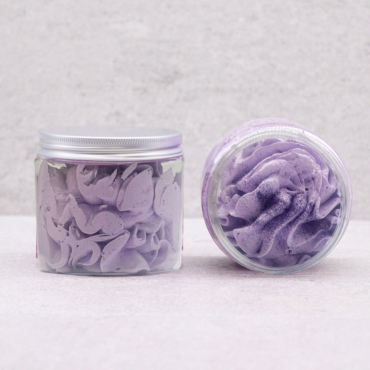 Two clear cosmetic jars containing sugar plum whipped soap one standing upright with a silver lid and one lying on its side, set against a light gray textured background.