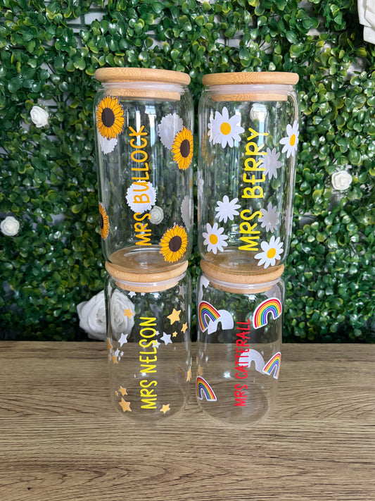 Four clear glasses with bamboo lids, decorated with daisies, sunflowers, rainbows, and personalised names, displayed on a green artificial grass background next to a wooden surface.