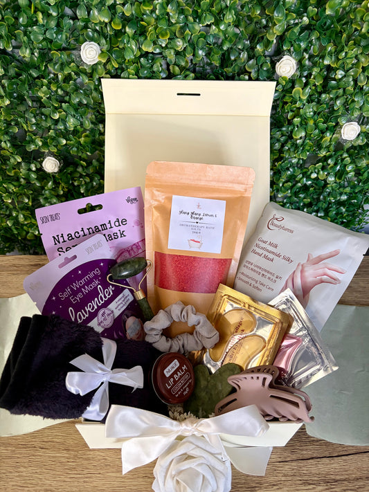 a curated self-care box including; facemask, hand mask, steam mask, eye patches, lip balm, face cloth, bath salts, hair scrunchie and clip, jade roller, gua sha, lip mask in a ivory gift box on a wooden surface with a green leafy backdrop.